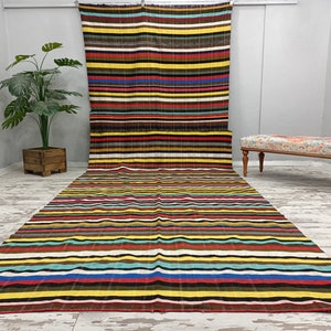 May include: A colorful striped rug with a variety of colors, including red, yellow, blue, and green. The rug is laid out on a white floor and a wooden bench is in the background.