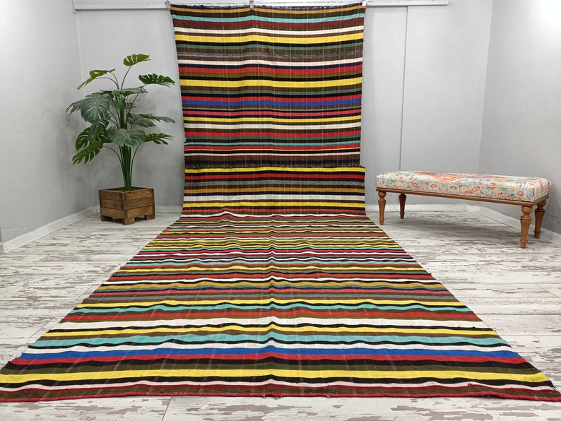 May include: A colorful striped rug with a variety of colors, including red, yellow, blue, and green. The rug is laid out on a white floor and a wooden bench is in the background.