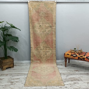 May include: A long, rectangular rug with a faded pink and beige colour scheme. The rug has a traditional Persian design with intricate floral patterns. It is hanging on a white wall in a room with a wooden floor.