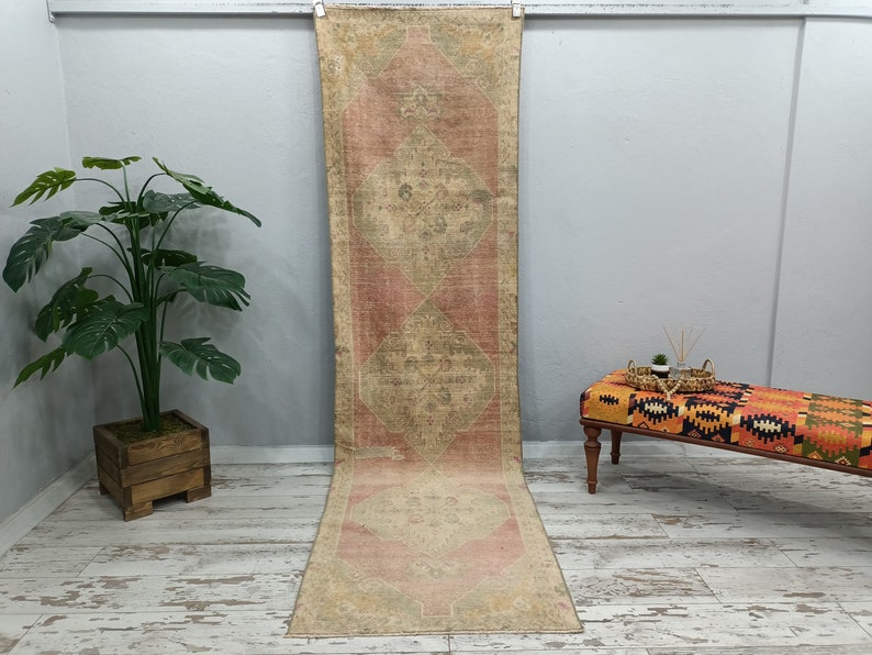May include: A long, rectangular rug with a faded pink and beige colour scheme. The rug has a traditional Persian design with intricate floral patterns. It is hanging on a white wall in a room with a wooden floor.