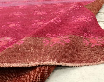Puede incluir: Una alfombra tejida de color rosa y marrón con un patrón floral. La alfombra tiene un borde de flecos y está doblada por la mitad.