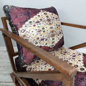 May include: A wooden armchair with a patterned cushion. The cushion features a burgundy background with a cream and gold floral design. The chair's wooden frame has a warm, brown finish, and the design is reminiscent of a vintage rug.