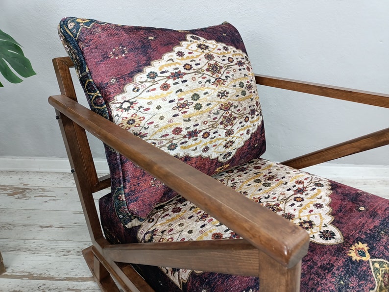 May include: A wooden armchair with a patterned cushion. The cushion features a burgundy background with a cream and gold floral design. The chair's wooden frame has a warm, brown finish, and the design is reminiscent of a vintage rug.