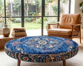 May include: A round ottoman featuring a blue fabric upholstery with a multi-colored intricate pattern, supported by four short wooden legs. The ottoman is positioned in front of a tan leather armchair and a stack of books on a wooden surface.