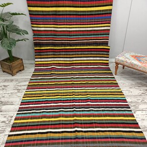 May include: A long, rectangular striped rug with a variety of colors, including red, yellow, blue, green, and brown. The rug is made of a woven fabric and has a fringed edge.