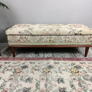 May include: A vintage storage bench with a floral patterned fabric top and wooden legs. The bench is sitting on a rug with a similar floral pattern.