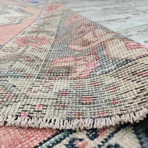 Pode incluir: Close-up da borda de um tapete vintage com franja. O tapete tem um fundo creme com padr&otilde;es rosa, azul e vermelho.