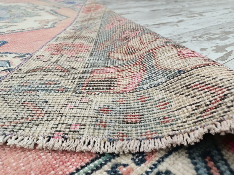Pode incluir: Close-up da borda de um tapete vintage com franja. O tapete tem um fundo creme com padr&otilde;es rosa, azul e vermelho.