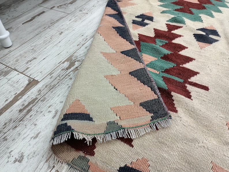K&ouml;nnte beinhalten: Ein beiger Teppich mit einem geometrischen Muster in Rosa-, T&uuml;rkis-, Marine- und Burgundert&ouml;nen. Der Teppich ist teilweise aufgerollt und zeigt die Unterseite mit einem Fransenrand. Das Design weist sich wiederholende Formen auf.
