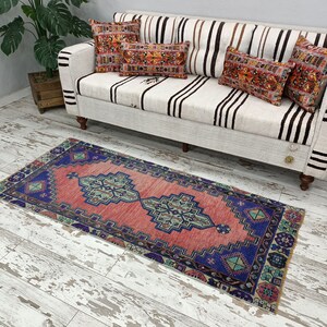 May include: A pink and blue patterned runner rug with a geometric design. The rug is placed in front of a white couch with black stripes.