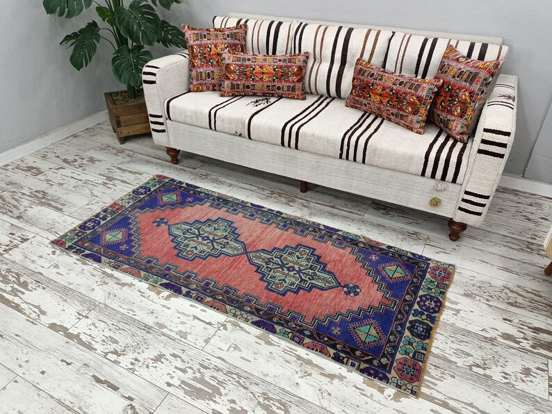 May include: A pink and blue patterned runner rug with a geometric design. The rug is placed in front of a white couch with black stripes.