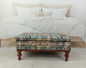 Op de afbeelding: Een kleurrijke, gepatroneerde opbergottoman met houten poten. De ottoman staat voor een wit bed met een wit deken en beige kussens.