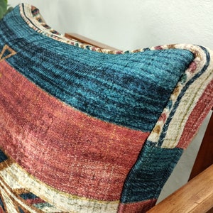 K&ouml;nnte beinhalten: Nahaufnahme eines gemusterten Kissens auf einem Holzstuhl. Das Kissen hat ein geometrisches Design mit t&uuml;rkisfarbenen, roten und beigen Streifen und Dreiecken. Der Stuhl hat eine dunkle Holzoberfl&auml;che.