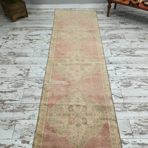 May include: A vintage runner rug with a faded pink and cream colour scheme. The rug has a traditional floral pattern with a diamond shape in the centre.