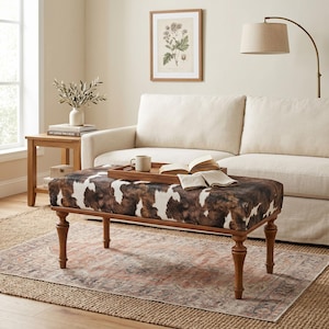 May include: A rectangular ottoman with a cowhide pattern in brown and white, set on four wooden legs. The ottoman is in a living room setting, with a sofa, side table, and rug visible.