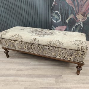 Op de afbeelding: Een beige en bruine opbergkist met houten poten. De kist staat op een lichte houten vloer.