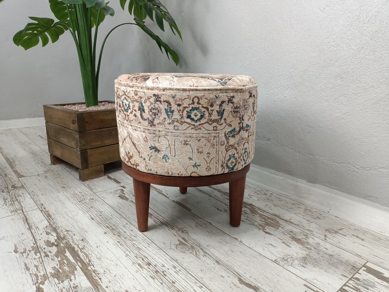 May include: A round ottoman with a beige fabric cover featuring a traditional Persian rug pattern. The ottoman has four wooden legs.
