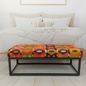 Op de afbeelding: Een rechthoekige poef met een kleurrijk patroon in oranje, rood, geel en groen. De poef staat op een zwart metalen frame. Het meubelstuk staat aan het voeteneinde van een bed met wit beddengoed.