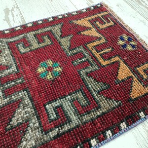 May include: A red, hand-woven rug with a geometric pattern in shades of gray, orange, and blue. The rug has a fringed edge.