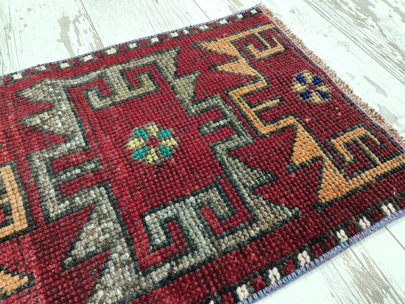May include: A red, hand-woven rug with a geometric pattern in shades of gray, orange, and blue. The rug has a fringed edge.
