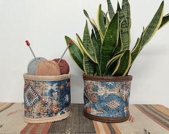May include: Two cylindrical fabric containers with a blue, orange, and brown patterned design. One container holds balls of yarn and knitting needles, while the other holds a green plant with yellow stripes.