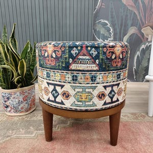 a round ottoman with a colorful, patterned design, placed on a pink rug. Next to the ottoman is a potted plant, and a white chair is visible in the background.