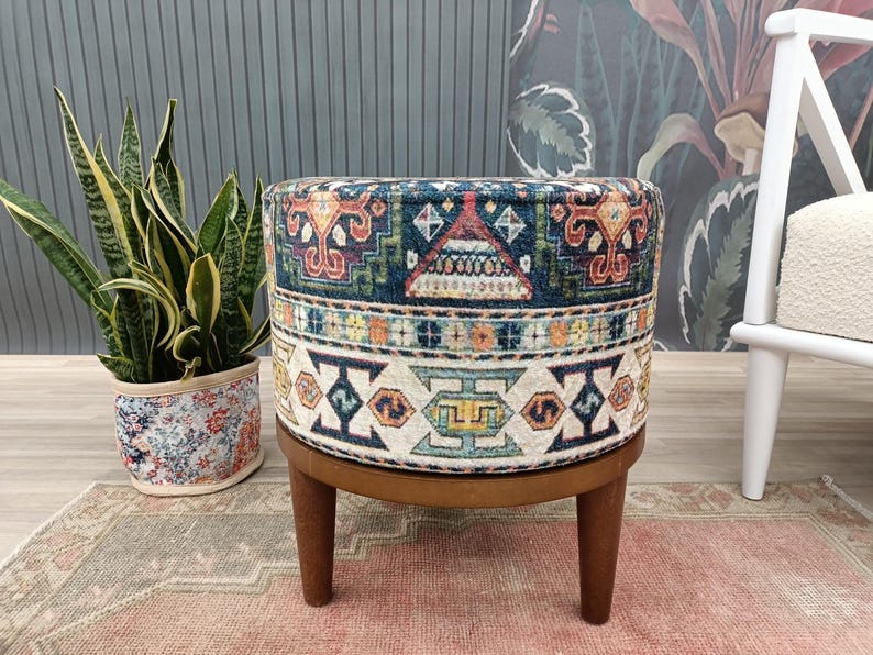 a round ottoman with a colorful, patterned design, placed on a pink rug. Next to the ottoman is a potted plant, and a white chair is visible in the background.