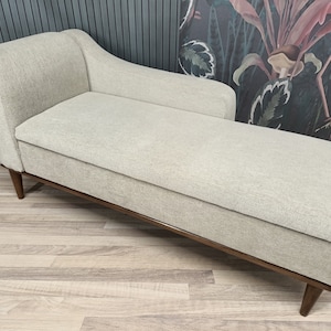 May include: A light gray chaise lounge with a wooden frame and legs. The chaise lounge is upholstered in a soft, textured fabric.