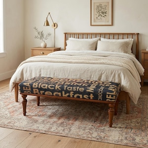 May include: A bedroom scene with a wooden bed frame and neutral bedding. A bench with a dark blue fabric top and wooden legs is at the foot of the bed. The fabric is printed with text. The rug has a patterned design.