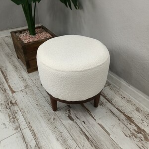 Puede incluir: Otomana redonda blanca con textura esponjosa y patas de madera. La otomana est&aacute; sobre un suelo de madera blanqueada.