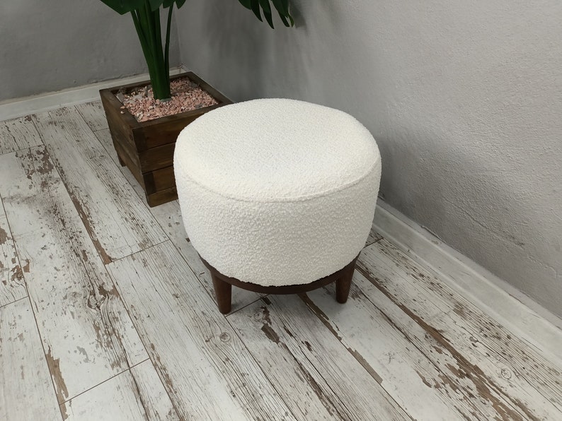 Puede incluir: Otomana redonda blanca con textura esponjosa y patas de madera. La otomana est&aacute; sobre un suelo de madera blanqueada.