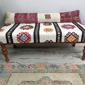 May include: A white and brown patterned ottoman with turned wooden legs. The ottoman has a long, red and white patterned pillow on top.
