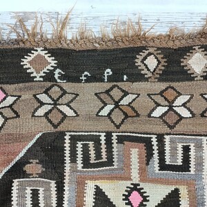 May include: Close-up of a brown and black rug with a geometric pattern. The rug has a fringe border and the letters "E F P" are woven into the rug.