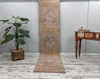 Könnte beinhalten: Ein langer, rechteckiger Teppich mit einem verblassten, Vintage-Look. Der Teppich hat einen hellbraunen Hintergrund mit einem sich wiederholenden Muster aus rosa und grauen Rauten. Der Teppich hängt an einer weißen Wand.
