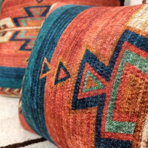 K&ouml;nnte beinhalten: Zwei Dekokissen mit einem s&uuml;dwestlichen Muster mit geometrischen Formen in Orange-, Rot-, Blau-, Gr&uuml;n- und Wei&szlig;t&ouml;nen.