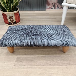 Peut inclure: Un repose-pieds rectangulaire avec un dessus à motifs bleus et gris et quatre pieds en bois courts et arrondis. Le repose-pieds est posé sur un sol en bois de couleur claire. Un pot de plante et une partie d'une chaise blanche sont visibles en arrière-plan.