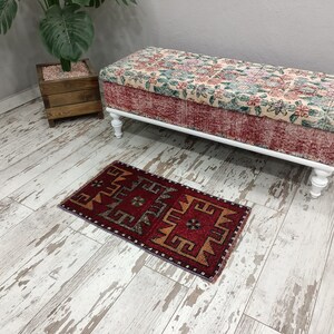 May include: A red, orange, and gray patterned rug with a geometric design sits on a whitewashed wood floor in front of a white upholstered bench with floral fabric.