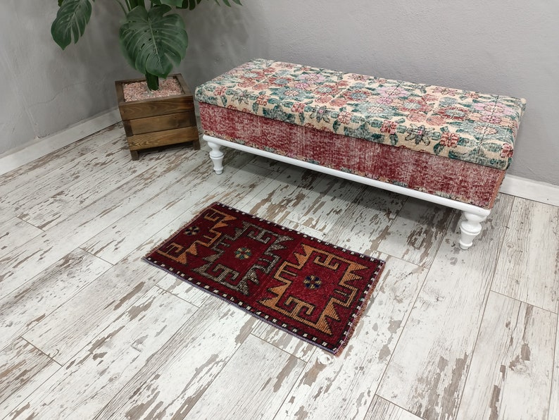 May include: A red, orange, and gray patterned rug with a geometric design sits on a whitewashed wood floor in front of a white upholstered bench with floral fabric.