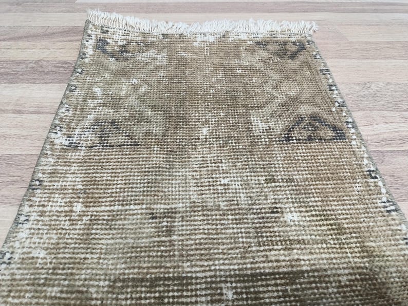 May include: A beige and brown woven rug with a fringed edge. The rug has a faded pattern with a geometric design.