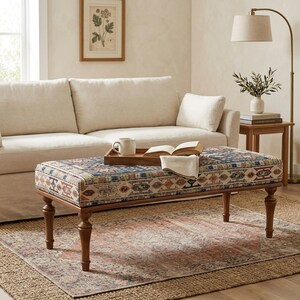 a cozy living room with a beige sofa, a wooden coffee table with a tray and a book, and a lamp on a side table. The room has a neutral color palette and a patterned rug on the floor.