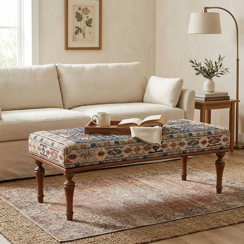 a cozy living room with a beige sofa, a wooden coffee table with a tray and a book, and a lamp on a side table. The room has a neutral color palette and a patterned rug on the floor.