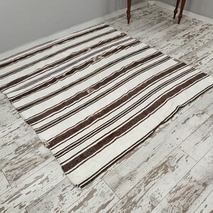 Puede incluir: Una alfombra rectangular con fondo blanco y rayas marrones oscuras espaciadas uniformemente. La alfombra est&aacute; hecha de un material tejido y tiene un dise&ntilde;o simple pero elegante. La alfombra est&aacute; colocada sobre un suelo de madera clara desgastada.