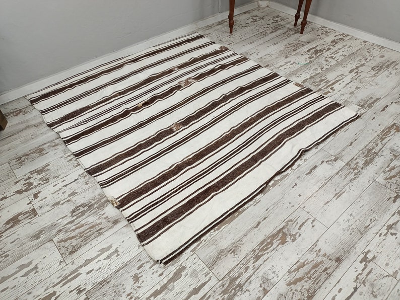 Puede incluir: Una alfombra rectangular con fondo blanco y rayas marrones oscuras espaciadas uniformemente. La alfombra est&aacute; hecha de un material tejido y tiene un dise&ntilde;o simple pero elegante. La alfombra est&aacute; colocada sobre un suelo de madera clara desgastada.