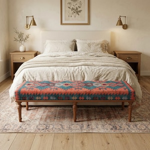 May include: A bedroom scene featuring a bed with a cream-colored headboard and bedding. A bench with a colorful geometric pattern in shades of red, teal, and blue sits at the foot of the bed. The bench has wooden legs. Two wooden nightstands flank the bed.