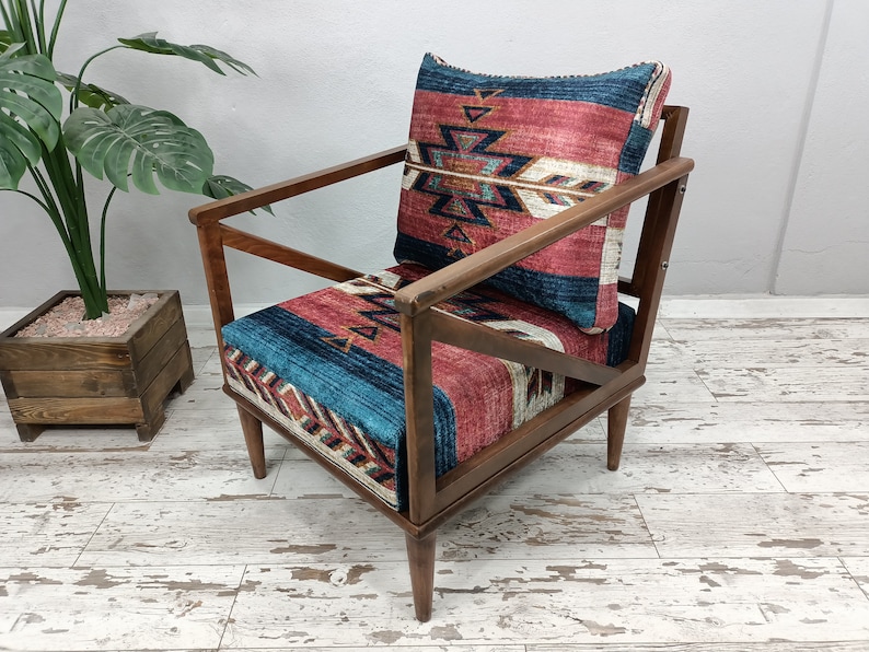 K&ouml;nnte beinhalten: Holzsessel mit geometrisch gemustertem Kissen und R&uuml;ckenlehne. Das Kissen hat ein Design in Rot-, Blau- und Wei&szlig;t&ouml;nen. Der Stuhl hat ein dunkles Holzgestell und abgewinkelte Beine, geeignet f&uuml;r ein Wohn- oder Schlafzimmer.
