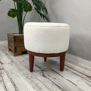 Puede incluir: Otomana redonda blanca con una cubierta de tela texturizada y cuatro patas de madera. La otomana est&aacute; sentada en un suelo de madera blanqueada.