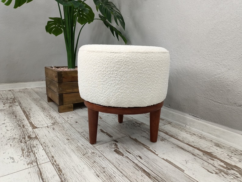 Puede incluir: Otomana redonda blanca con una cubierta de tela texturizada y cuatro patas de madera. La otomana est&aacute; sentada en un suelo de madera blanqueada.