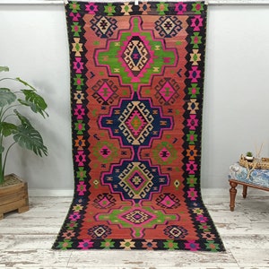 Puede incluir: Una alfombra kilim turca tradicional con un patr&oacute;n geom&eacute;trico en tonos de rosa, verde, azul y negro. La alfombra tiene un fondo rojo y un borde negro.
