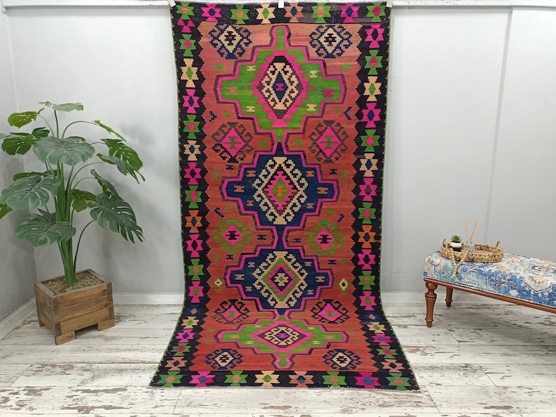 Puede incluir: Una alfombra kilim turca tradicional con un patr&oacute;n geom&eacute;trico en tonos de rosa, verde, azul y negro. La alfombra tiene un fondo rojo y un borde negro.