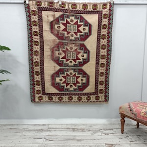 May include: A vintage Turkish rug with a cream background and three red, blue, and green geometric patterns. The rug is hanging on a white wall.